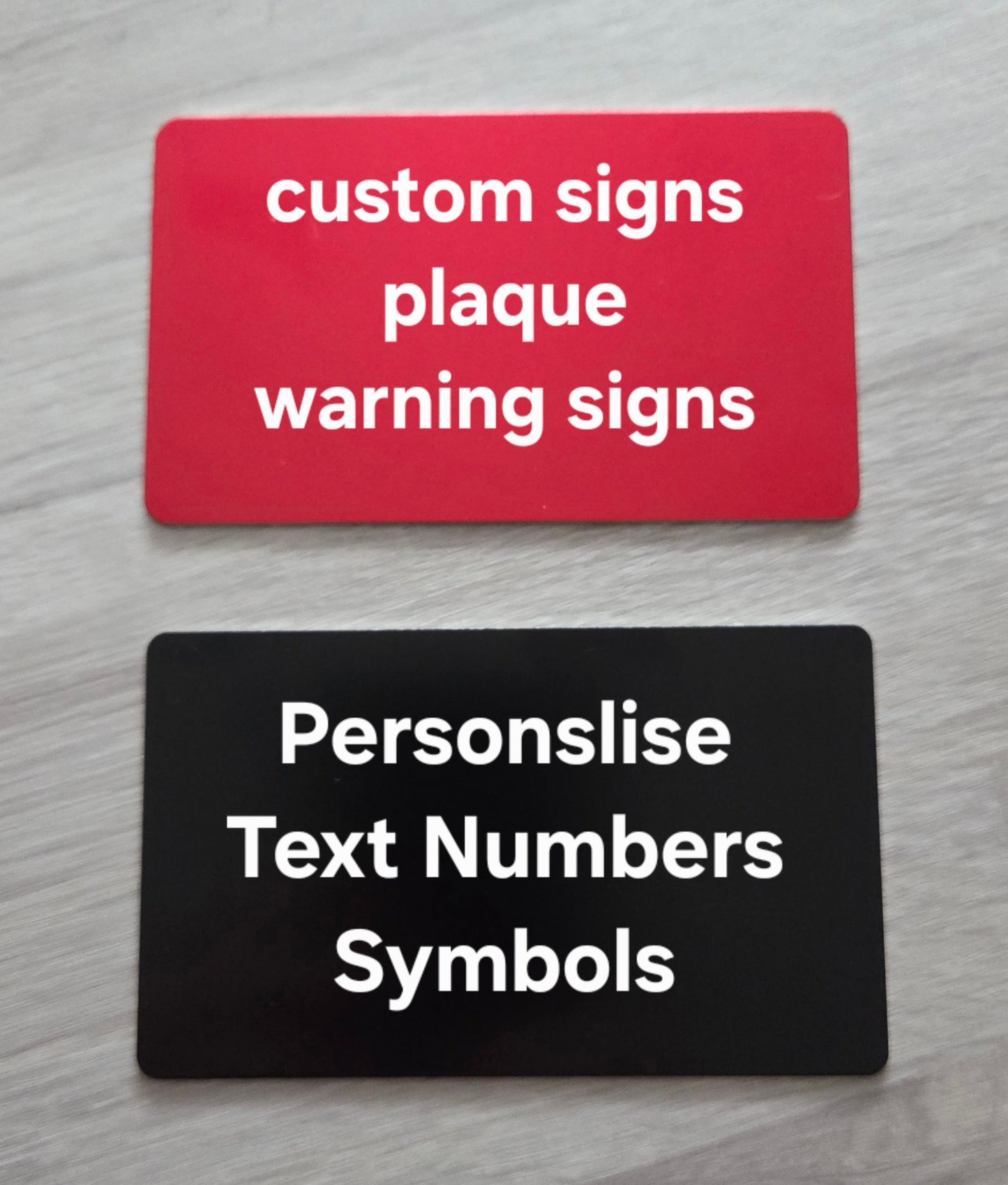 Two customizable signs on a gray surface, one red with white text and one black with white text.